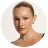 A young woman with light skin and pulled-back hair is shown in a close-up portrait against a plain, light background, wearing a sleeveless top and minimal makeup.
