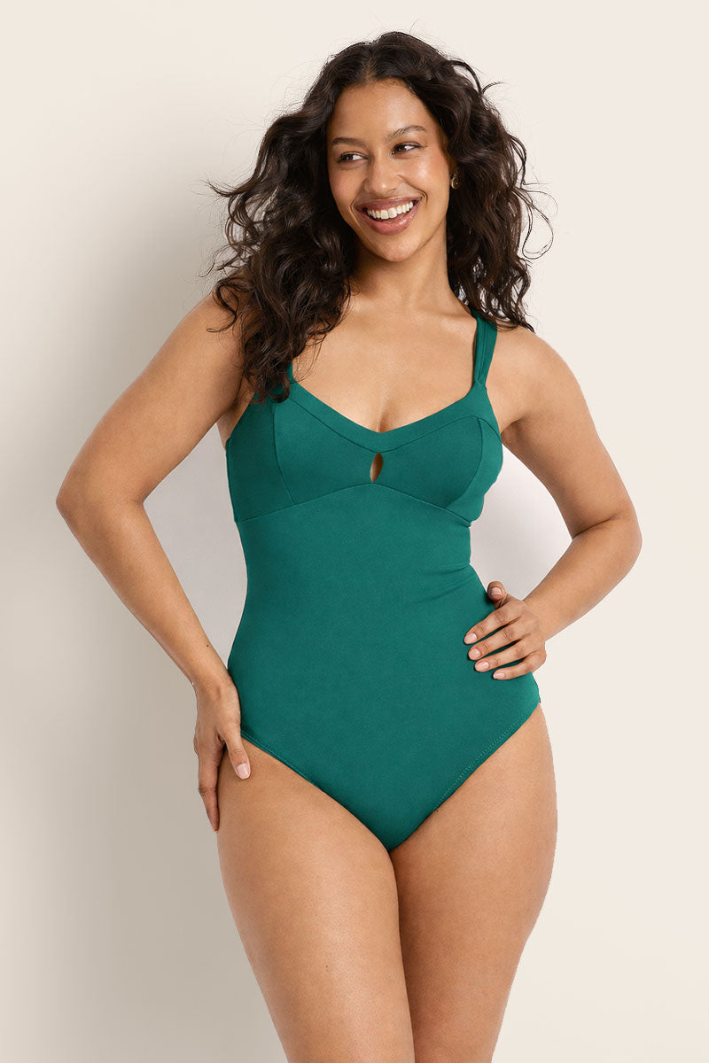 A woman with long wavy hair smiles while modeling the Fearless au Magic Suit - Teal / Standard, a one-piece swimsuit made from recycled fabric with wide straps, chest keyhole cutout, and built-in bust support against a plain backdrop.