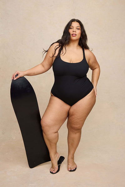 A plus-size woman modeling a black one-piece swimsuit, holding a black surfboard against a textured beige studio backdrop.