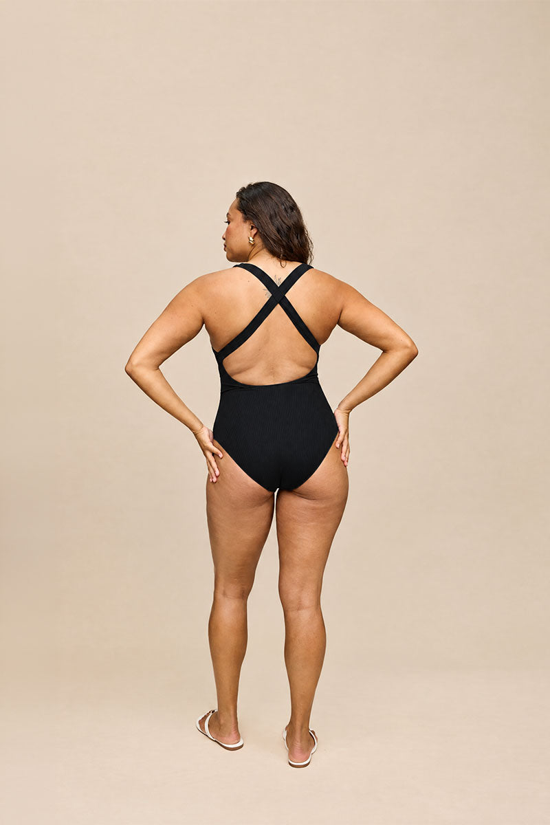 A woman modeling a black one-piece swimsuit from the back, showing the crisscross back straps, against a solid beige studio background.