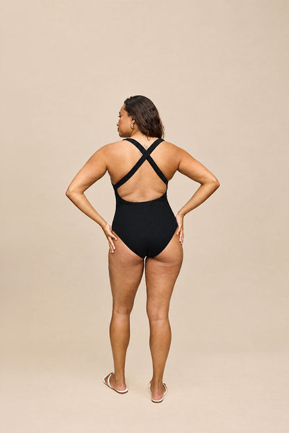 A woman modeling a black one-piece swimsuit from the back, showing the crisscross back straps, against a solid beige studio background.