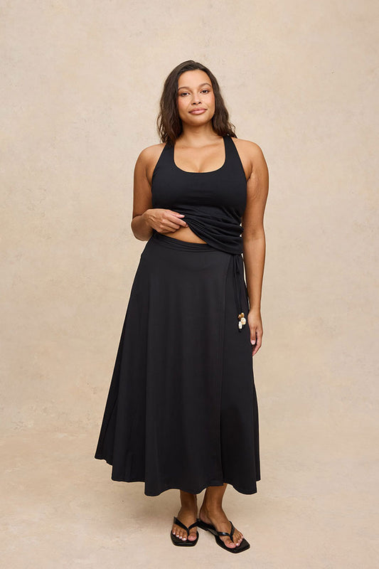 A woman modeling a black tank and skirt with a hidden shorts, holding out the side drape of the skirt against a textured beige studio backdrop.