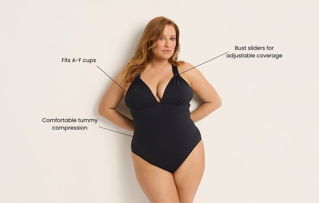 A woman models a black one-piece swimsuit. Text points out features: fits A–F cups, comfortable tummy compression, and bust sliders for adjustable coverage. She stands against a plain, light background with arms behind her back.