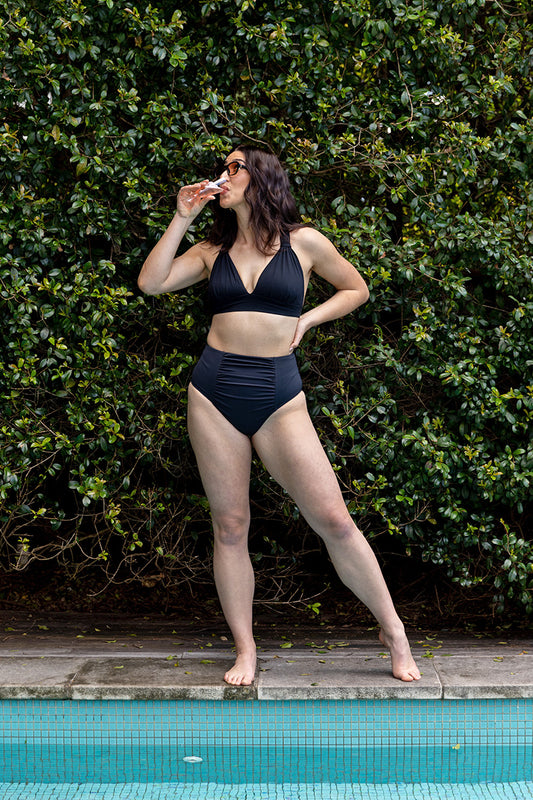 Wearing the All Day Bikini Top-Matte Black by Fearless au, a woman stands confidently by the pool with a drink in hand, sunglasses on, and one hand on her hip amid lush greenery.