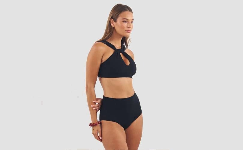 A woman wearing a stylish black two-piece swimsuit with a high-waisted bottom and a halter-style top featuring a keyhole cutout, standing against a plain light background.
