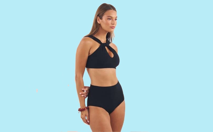 A woman stands against a light blue background, wearing a black two-piece swimsuit with high-waisted bottoms and a halter-style top featuring a keyhole cutout at the chest.