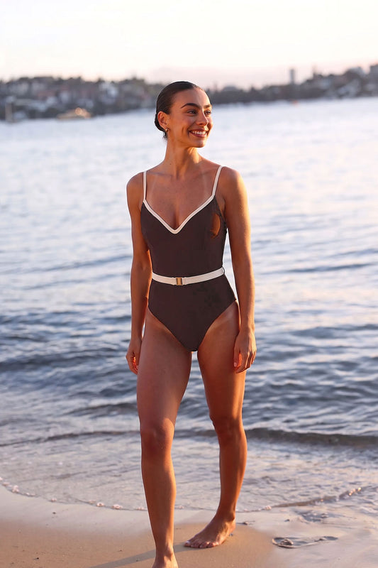 Woman in a brown swimsuit standing on a beach with water and skyline in the background
