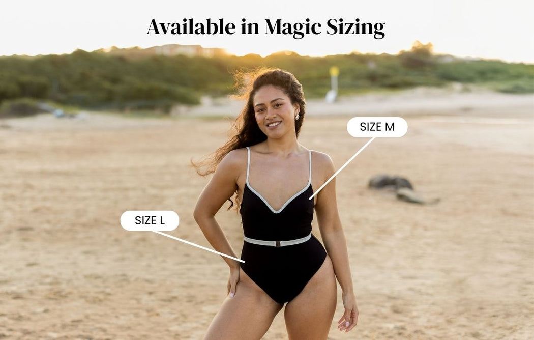 Woman in a black one-piece swimsuit stands on a sandy beach at sunset. Text reads Available in Magic Sizing. Arrows point to the suit labeled Size M and Size L. Greenery and hills are in the background.