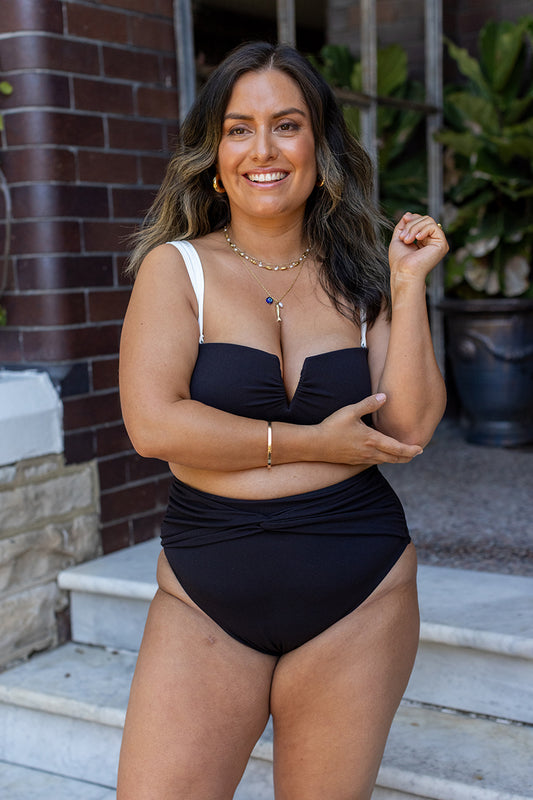 A woman with long dark hair smiles outdoors on steps, wearing the Fearless au Icon Bikini Top with adjustable straps. One arm crosses her body; she's surrounded by brick walls, greenery, and a large potted plant in the background.