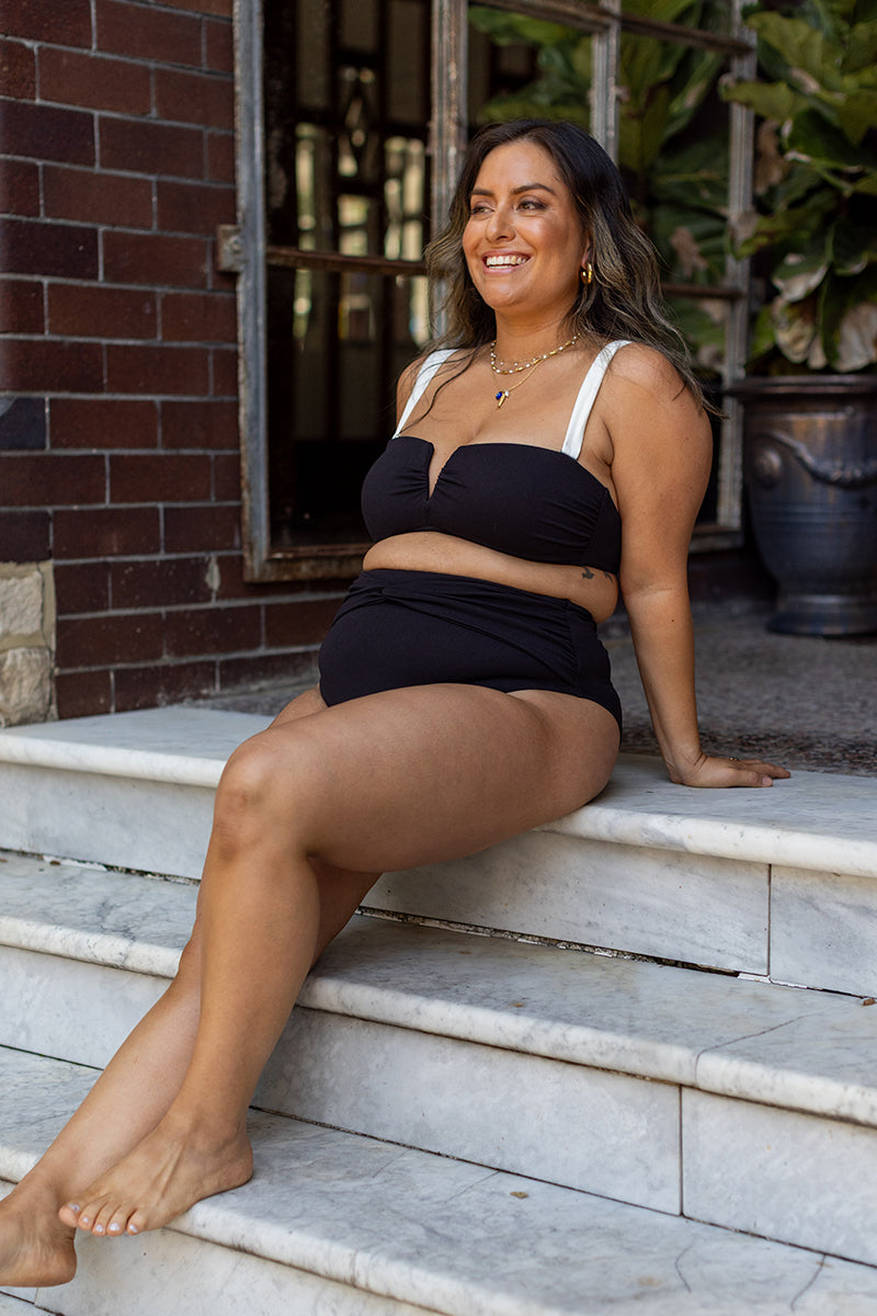 A smiling woman wearing a black and white bikini sits on marble steps outside, leaning back on one arm, with a brick wall and large potted plant in the background.