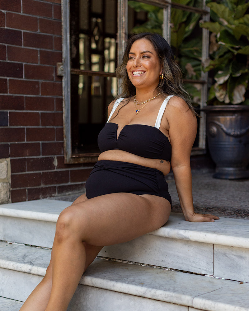 A smiling woman sits on marble steps outdoors, wearing a black bikini with white straps. She has long, wavy hair and is accessorized with layered necklaces. There are plants and a brick building in the background.