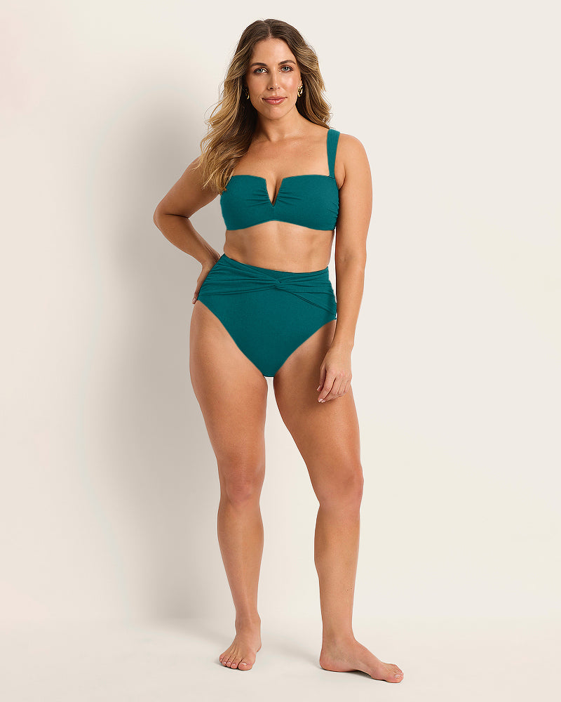 A woman with long, wavy hair stands barefoot against a neutral background, wearing Fearless au’s limited edition Icon Bikini Bottom in teal with a V-cut bandeau top, radiating confidence and relaxed style.