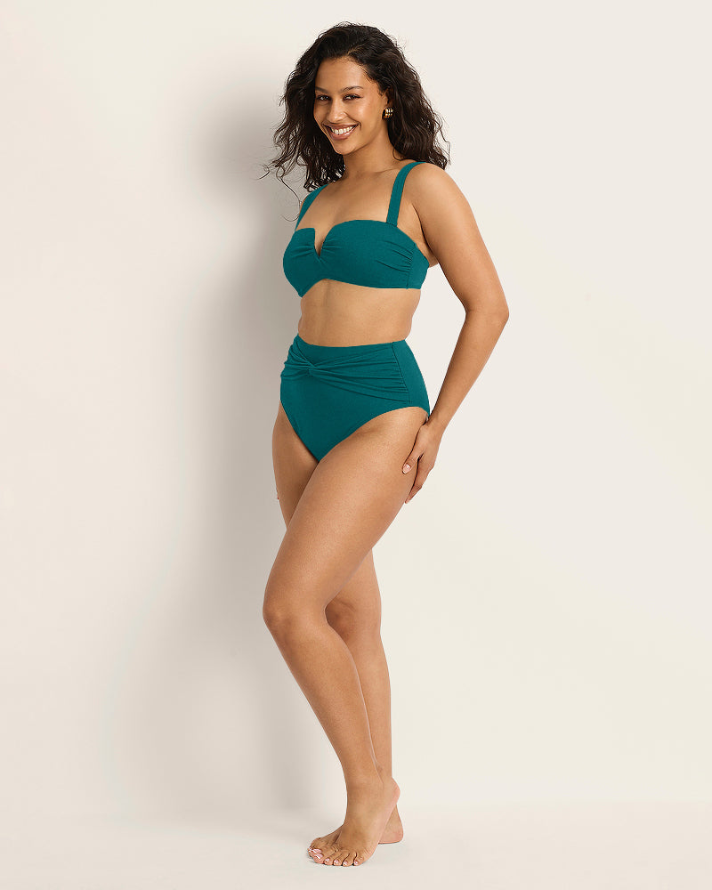 A woman with curly hair smiles while posing in the Fearless au Icon Bikini Bottom in teal, crafted from recycled crinkle fabric with a structured top. She stands barefoot against a plain light background, showcasing this limited edition style.
