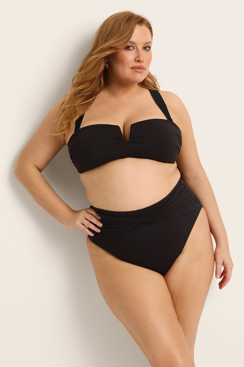 A woman with long, wavy hair poses confidently in Fearless au's Icon Bikini Top, a black two-piece made from recycled crinkle fabric. The structured top has wide straps and the high-waisted bottoms feature chic draped detailing.