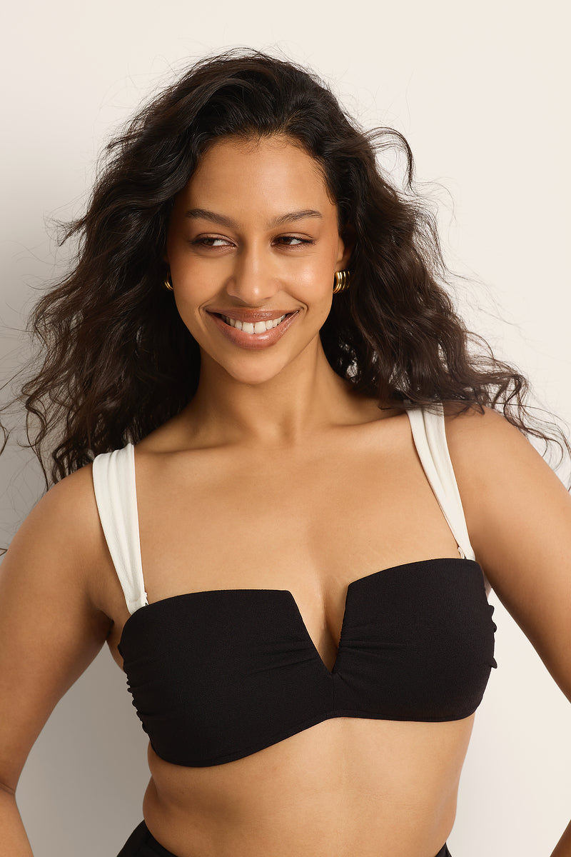A woman with long wavy hair smiles while wearing the Fearless au Icon Bikini Top in black, featuring adjustable straps and gold hoop earrings, set against a plain light-colored background.