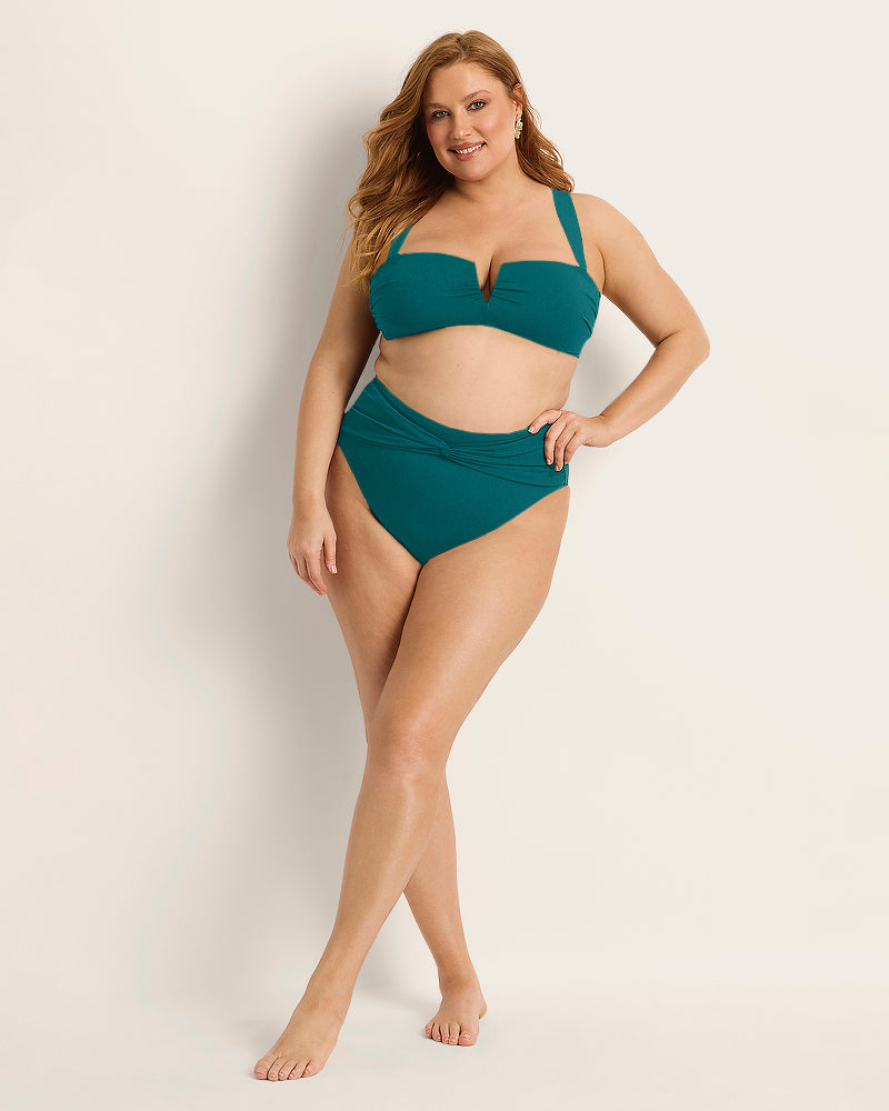A woman with long, light brown hair wears the Fearless au Icon Bikini Bottom, a limited edition teal high-waisted piece crafted from recycled crinkle fabric, standing barefoot and smiling against a plain beige background.