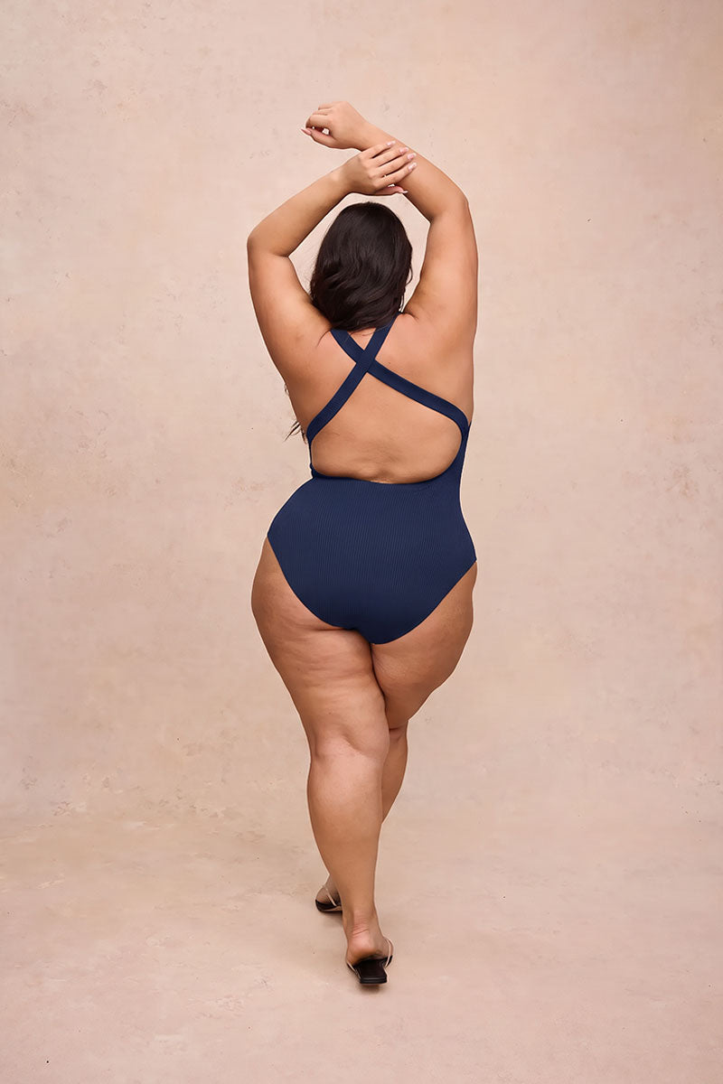 A woman is shown from behind wearing a navy blue one-piece swimsuit with crisscross back straps, standing against a textured beige studio backdrop.