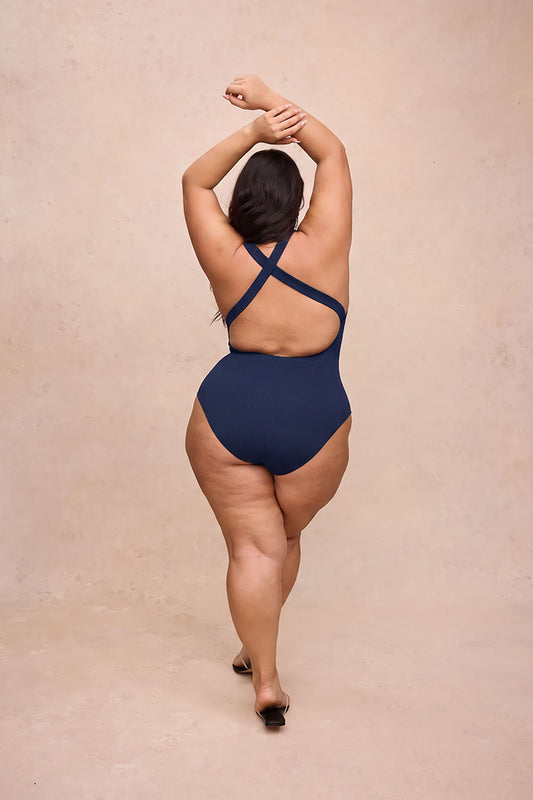 A woman is shown from behind wearing a navy blue one-piece swimsuit with crisscross back straps, standing against a textured beige studio backdrop.