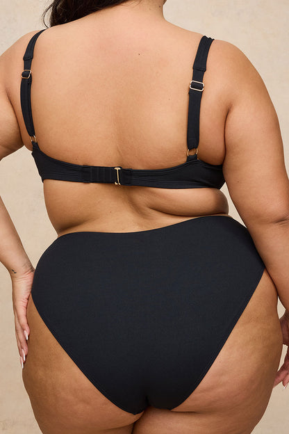 Rear view of a 2X model wearing a black two-piece swimsuit with adjustable back straps and gold-tone hardware, set against a neutral beige background.