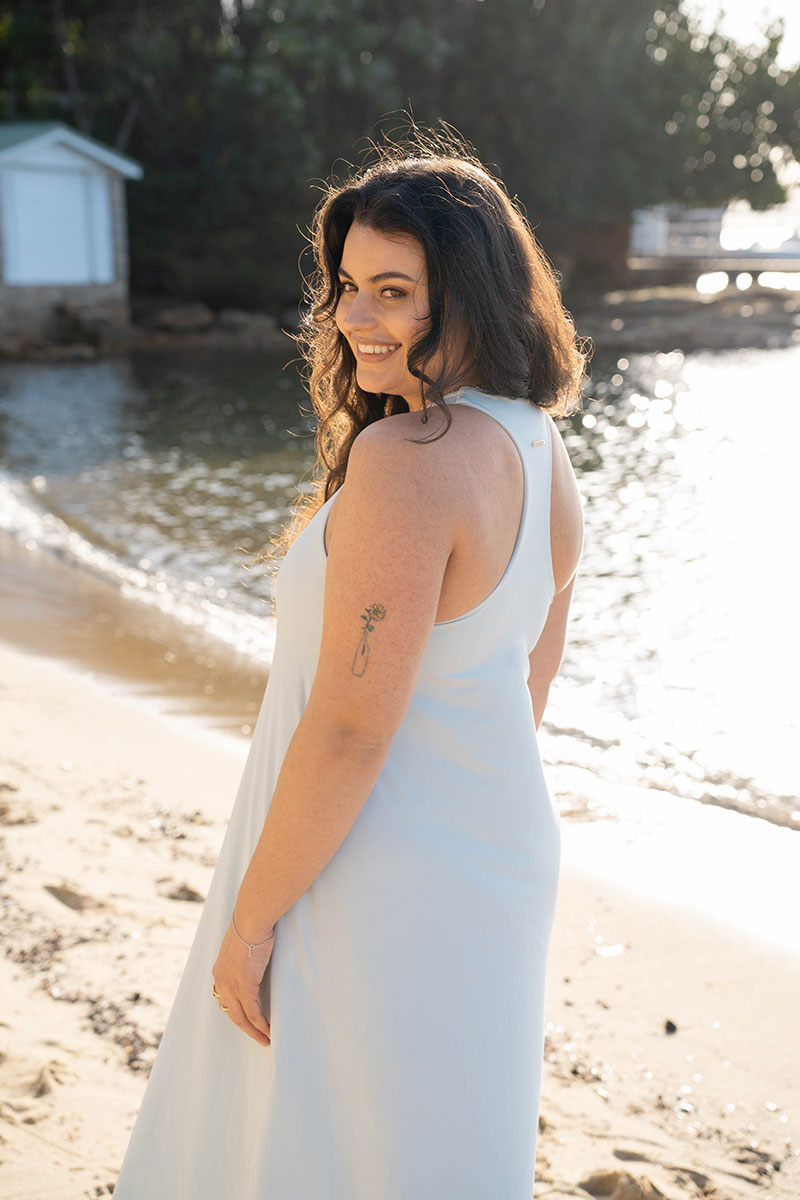 A woman with long dark hair stands on a sandy beach wearing a sleeveless white dress, looking back over her shoulder. The background shows water, trees, and a small white structure near the shore.