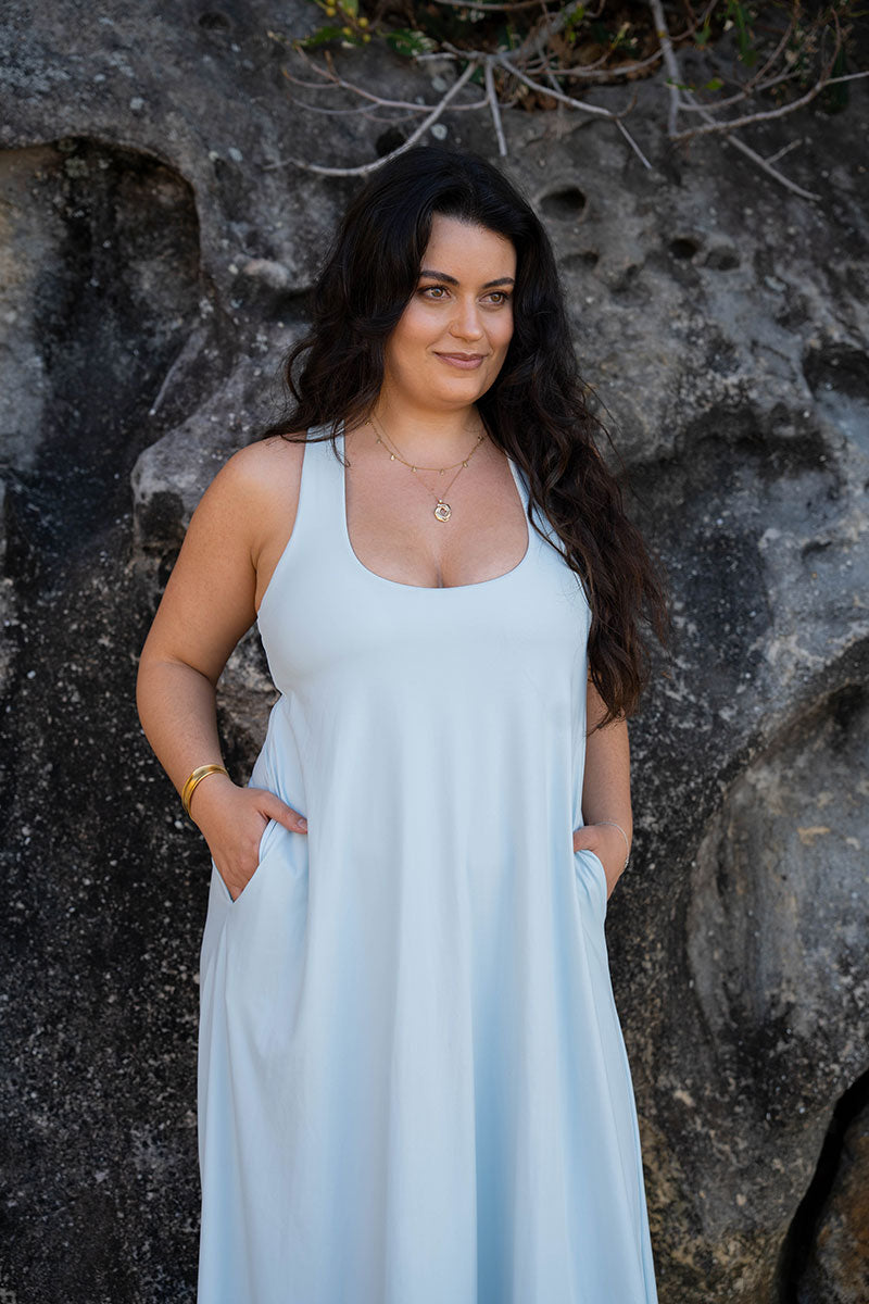 A woman with long dark hair wearing a light blue sleeveless maxi dress with side pockets, standing in front of a textured gray rock wall.
