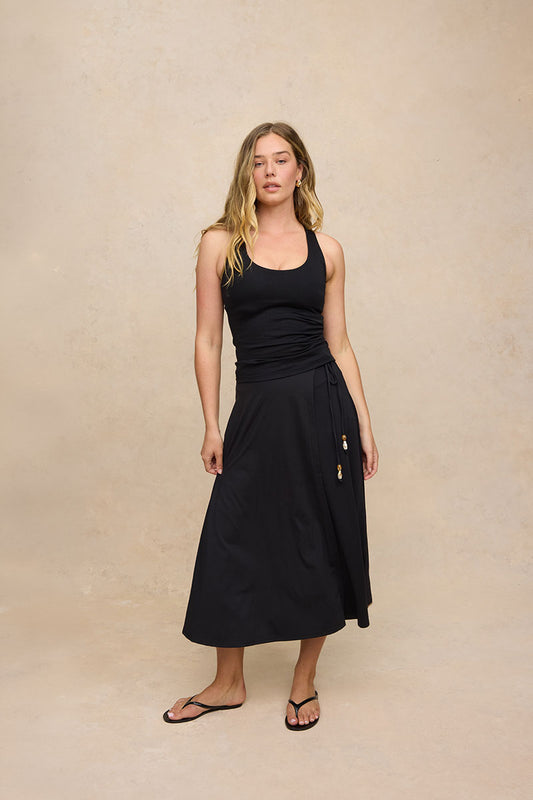 A woman modeling a black sleeveless midi dress with a ruched bodice and a drawstring waist, paired with black flip-flop sandals, against a textured beige studio backdrop.