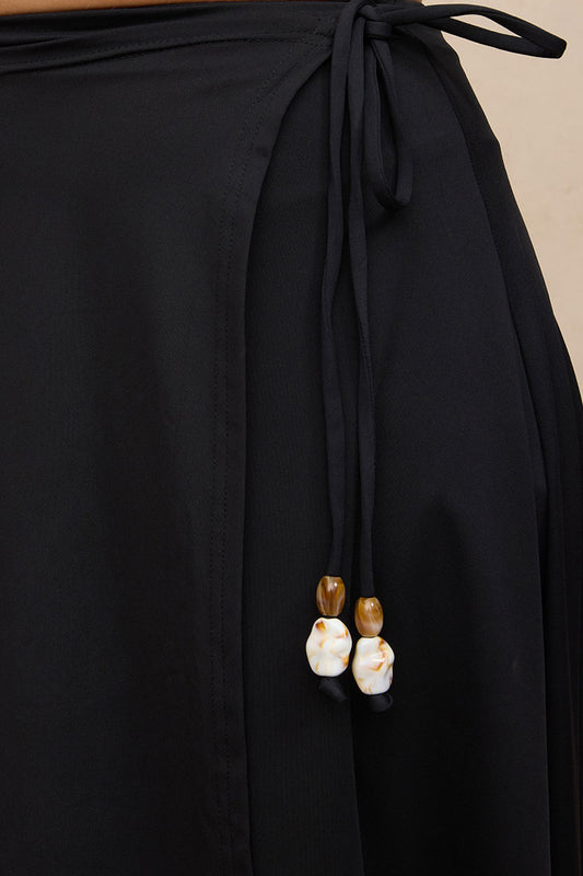 Close-up of a black wrap-style garment with a drawstring waist and two dangling bead accents, shown against a plain light beige background.