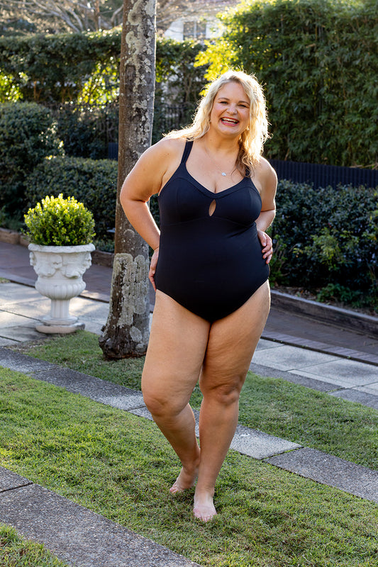 A smiling woman stands on grass, wearing the Fearless au Magic Suit - Matte Black / Standard. Lush greenery and a potted plant are visible in the bright, sunny background.