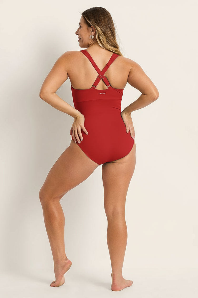 A woman stands barefoot on a light surface, facing away, smiling over her left shoulder while wearing the Fearless au Magic Suit - Standard: a red supportive swimsuit with crisscrossed straps and built-in bust support.
