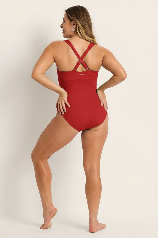 A woman stands barefoot on a light surface, facing away, smiling over her left shoulder while wearing the Fearless au Magic Suit - Standard: a red supportive swimsuit with crisscrossed straps and built-in bust support.
