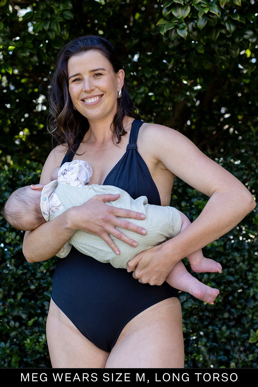 A woman wearing the Fearless au All Day One Piece - Standard in black (size M, long torso) stands outdoors smiling and holding a baby, with greenery in the background.