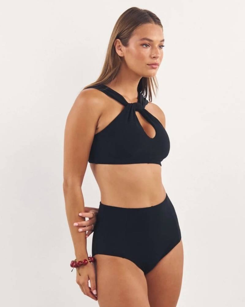 A woman stands against a plain background wearing a stylish black bikini with a keyhole cutout top and high-waisted bottoms. She has straight, light brown hair and is accessorized with a red beaded bracelet.