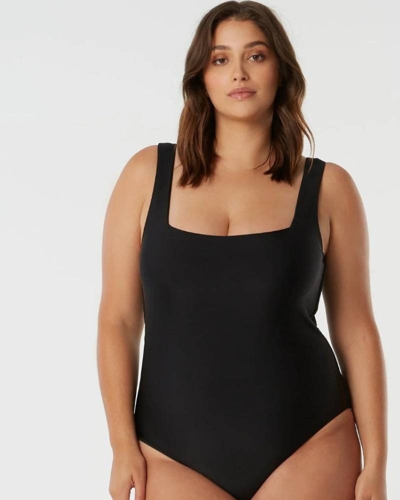 A woman with medium-length brown hair wears a simple black one-piece swimsuit and stands against a plain light background, looking confidently at the camera.