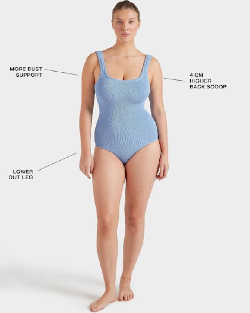 A woman stands facing forward wearing a light blue one-piece swimsuit. Text highlights features: more bust support, lower cut leg, and a 4 cm higher back scoop. The background is plain white.